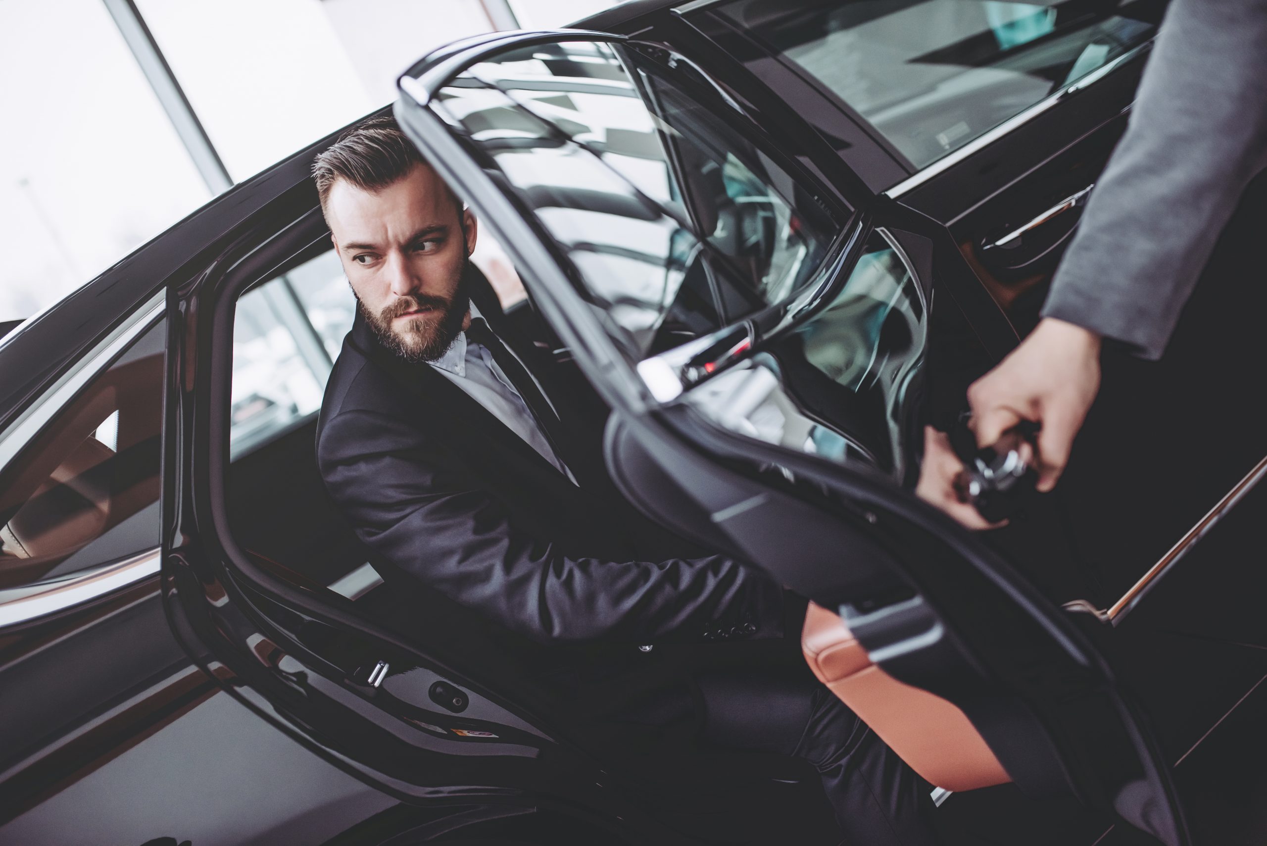 Young handsome businessman is sitting in luxury car. Serious bearded man in suit is coming out while driver is opening door.