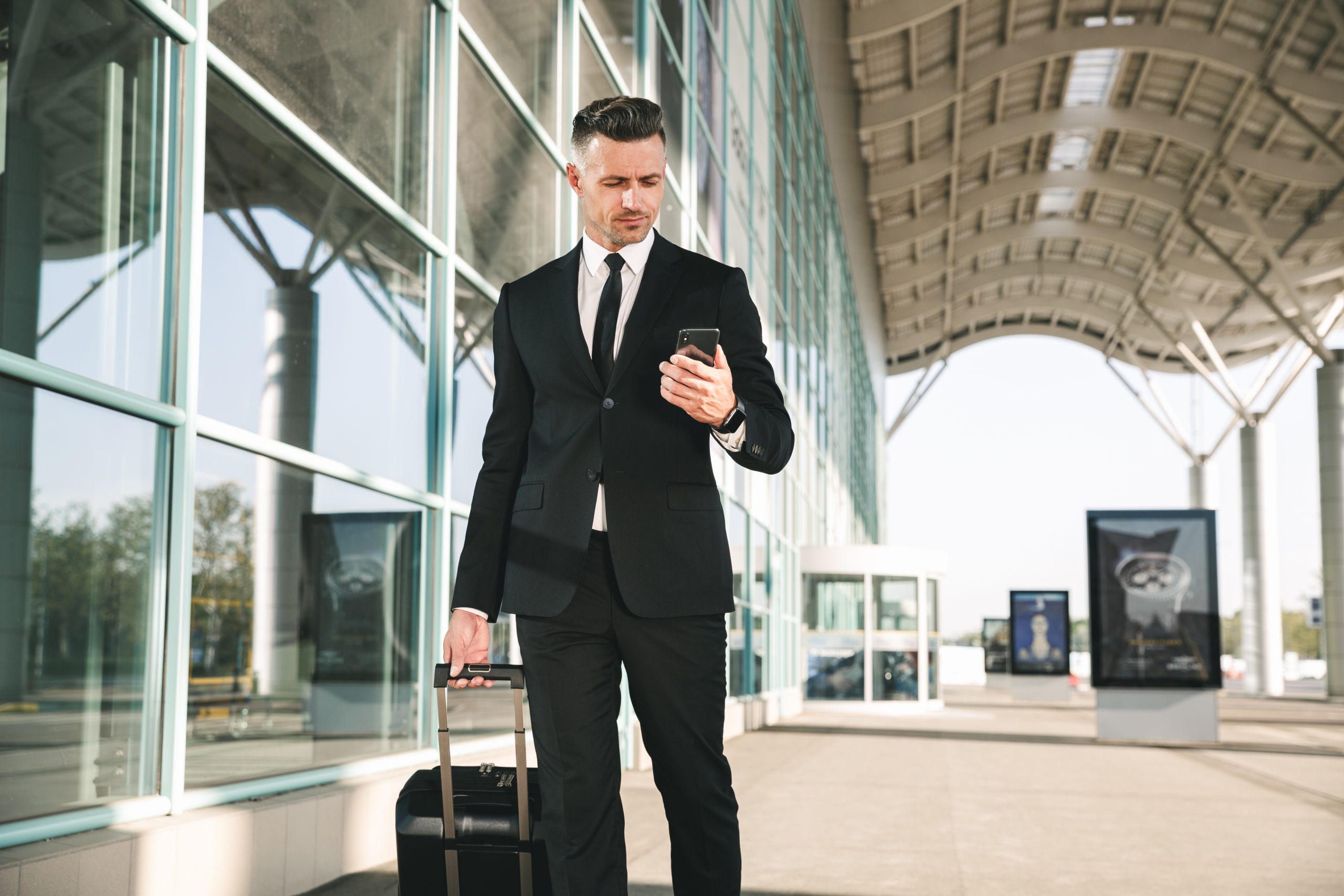 Handsome businessman dressed in suit walking with a suitcase outside airport terminal and looking at mobile phone